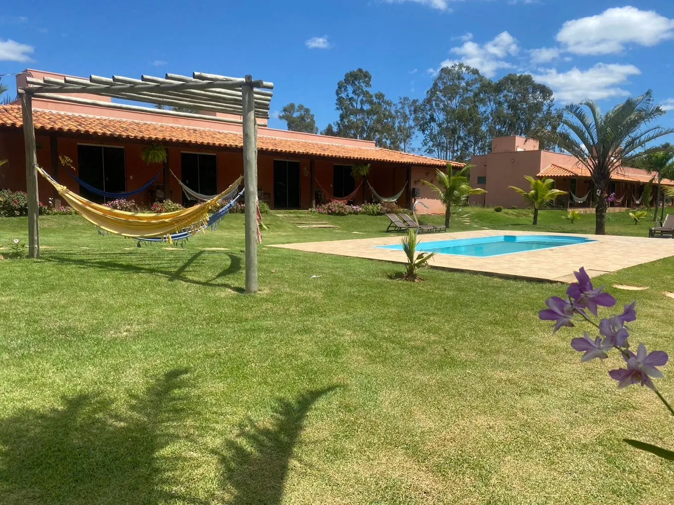 Jardim ensolarado com redes e piscina da pousada